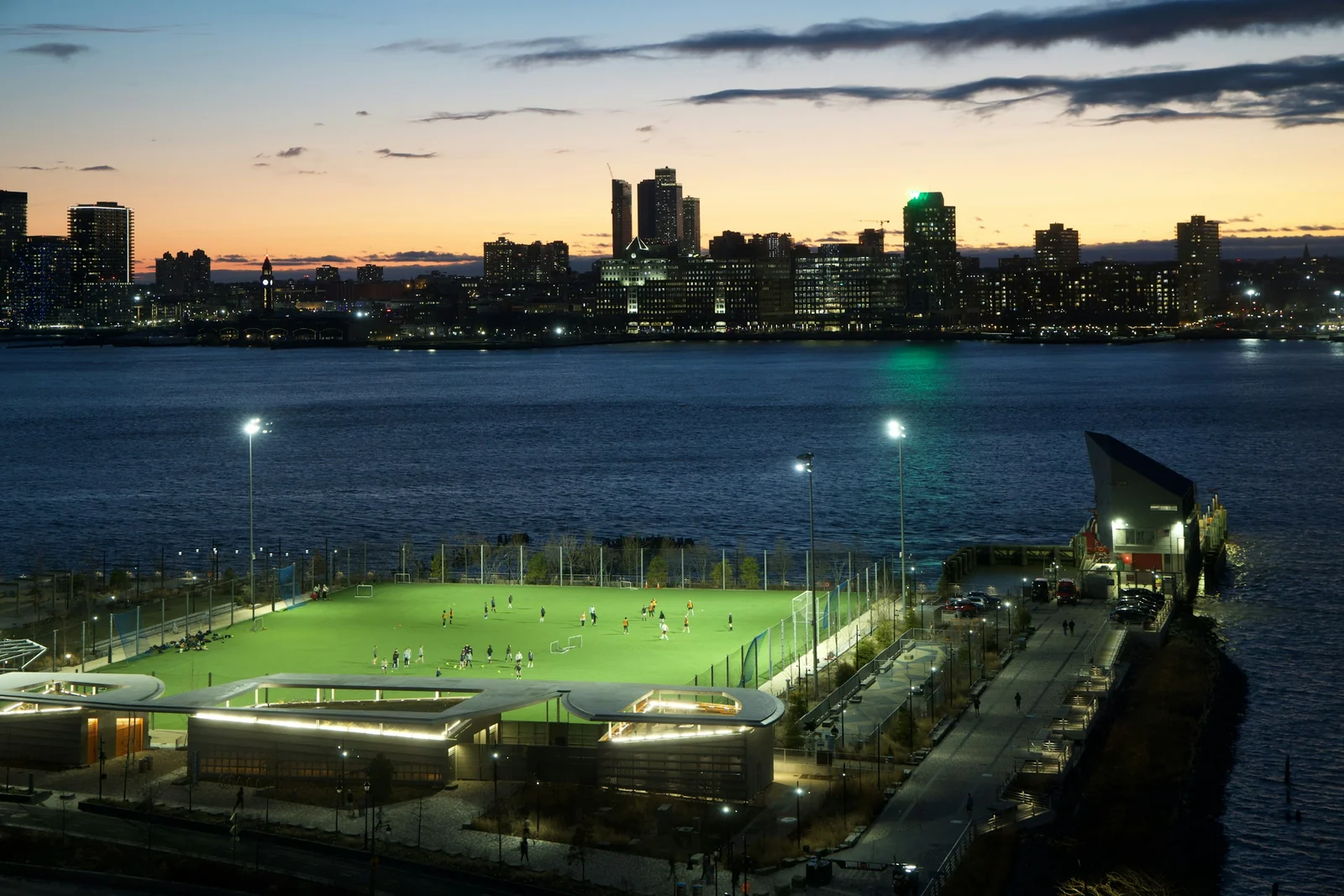 Players use a floodlit soccer field beside the Hudson River, with New Jersey buildings across the water at dusk.