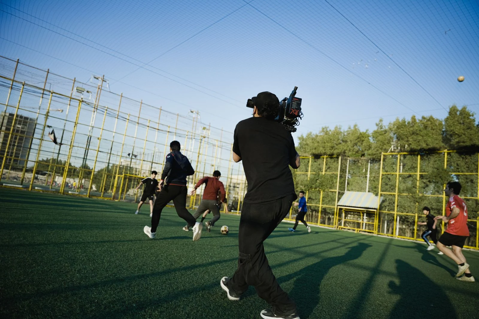 A camera operator films players on an enclosed outdoor soccer field.